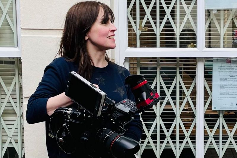 Оператор-постановщик Эмма Эдвардс смотрит влево, удерживая в руках камеру Canon во время съемок рядом со зданием с большими окнами.