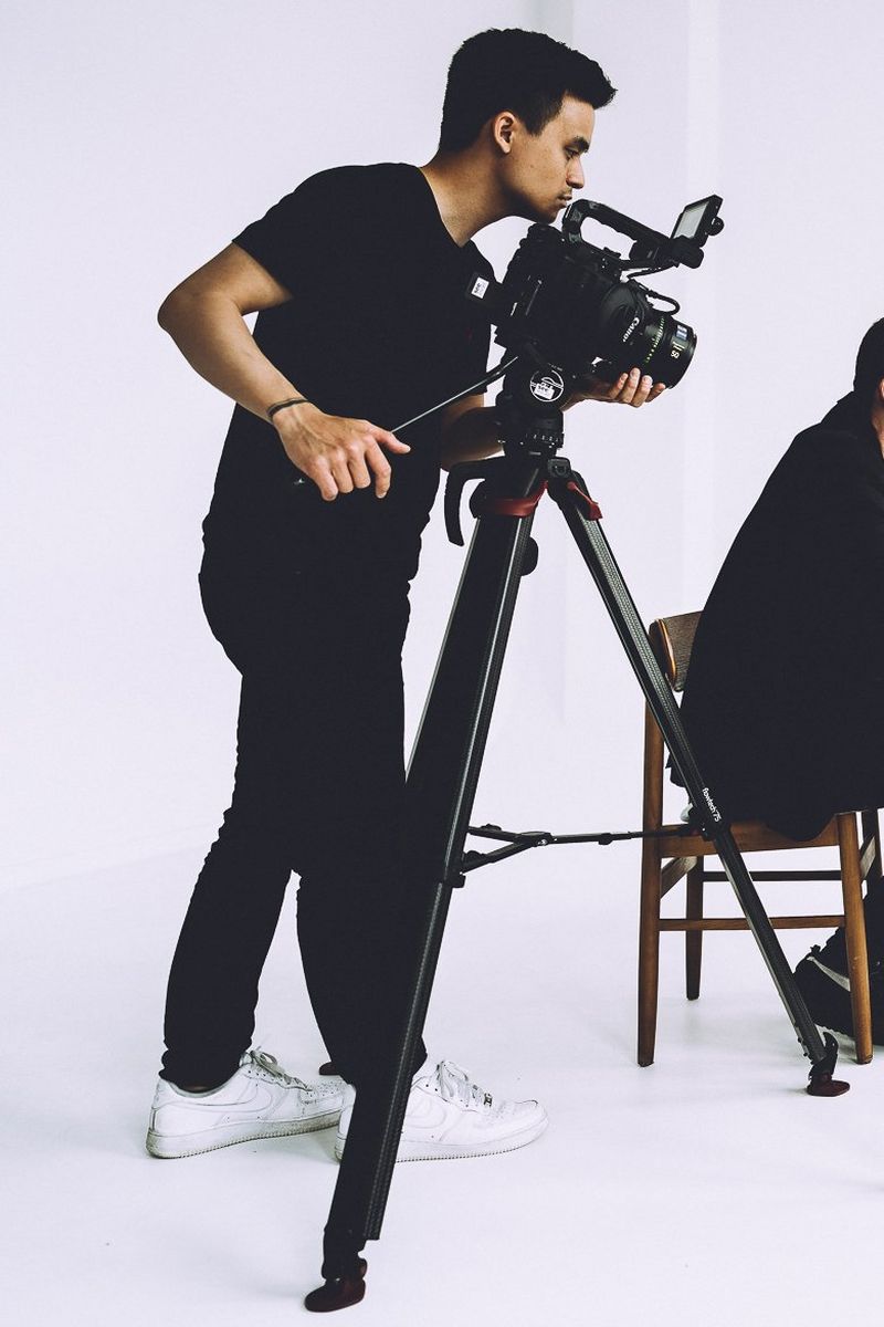 Видеограф Денни Фэн в повседневной одежде и белых кроссовках снимает на камеру Canon на светло-пурпурном фоне.