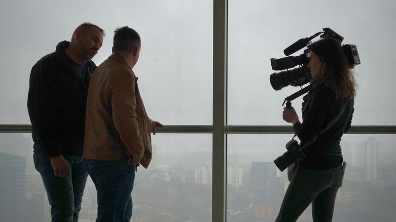 Оператор-постановщик Эмма Эдвардс снимает видео с диалогом двух мужчин у окна во французском стиле. Один из них держится за перила и смотрит на покрытый туманной дымкой город.