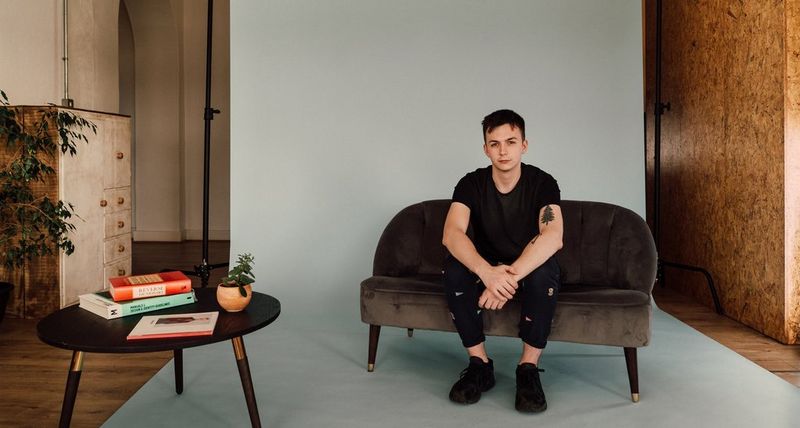 Создатель контента и маркетинговый директор Харри Ситон сидит на небольшом коричневом диване, находясь на съемочной площадке, повторяющей интерьер гостиной.