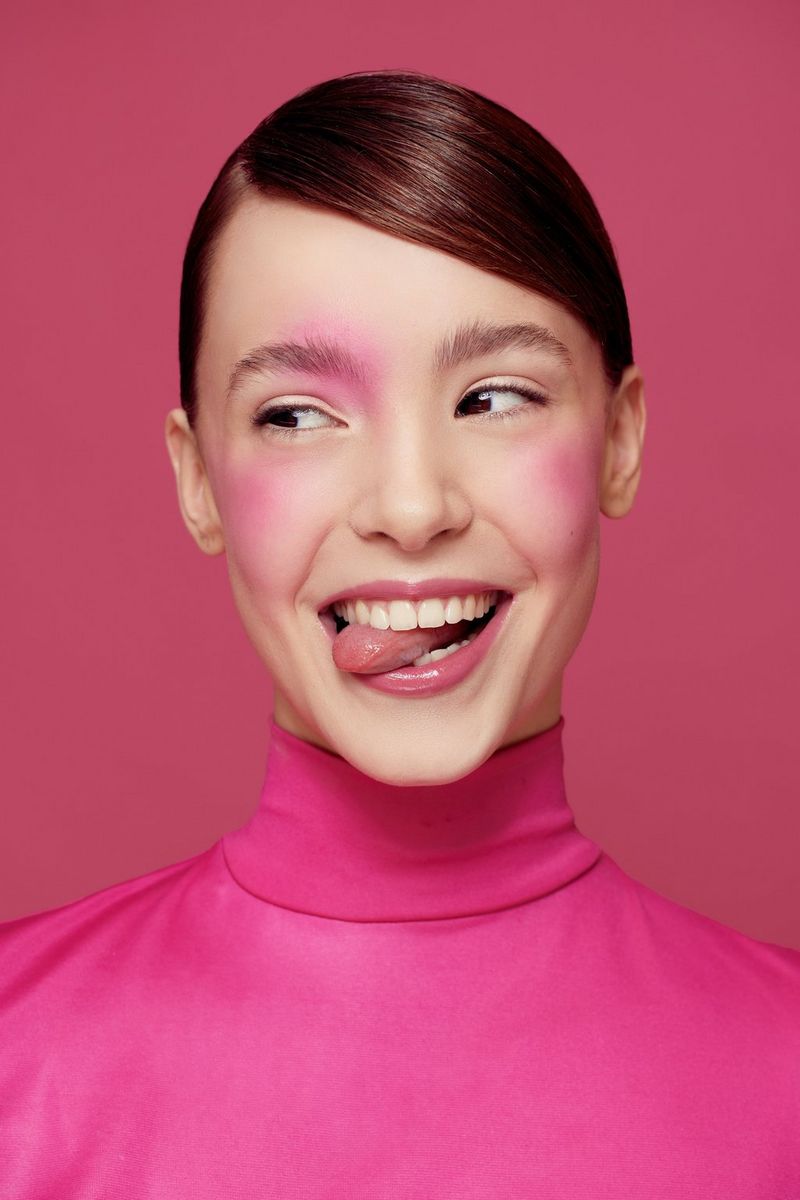 Una foto scattata da Evely Duis del primo piano di una modella vestita di rosa, su uno sfondo rosa, che mostra la lingua su un lato della bocca.