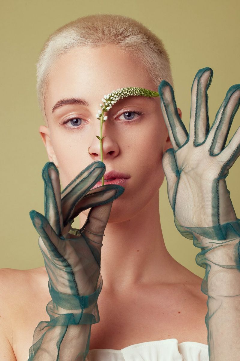 Uno scatto di Evely Duis che mostra un primo piano di una modella con i capelli rasati, le cui mani guantate reggono un fiore davanti al viso, curvandolo affinché segua la linea del sopracciglio.