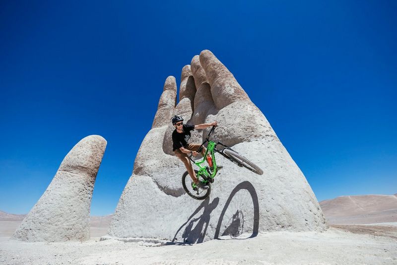A mountain biker performs a jumps on a stone statue built in the shape of a large hand, which is rising out of dusty and sparse terrain. 