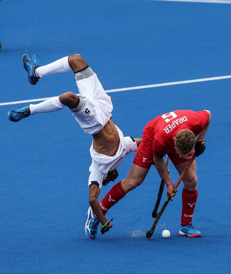 A hockey player in red has taken the ball from a hockey player in white, who has been thrown in the air and upside down by the tackle.