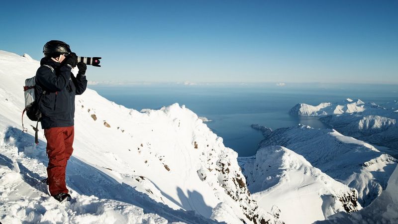 A photographer wearing winter clothing and carrying a backpack stands on a snow-covered mountainside holding a Canon camera. © Richard Walch