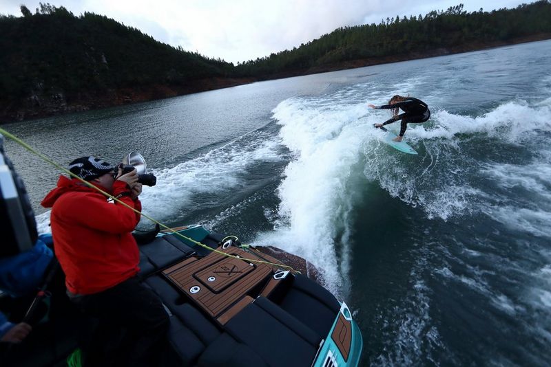 Een man in een rode hoodie en een zwarte pet maakt vanaf de achterkant van een snelle boot een foto van een wakesurfende vrouw.