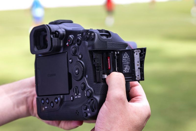 A user holds a Canon EOS R1 with its card slot cover open, showing two memory cards inserted.