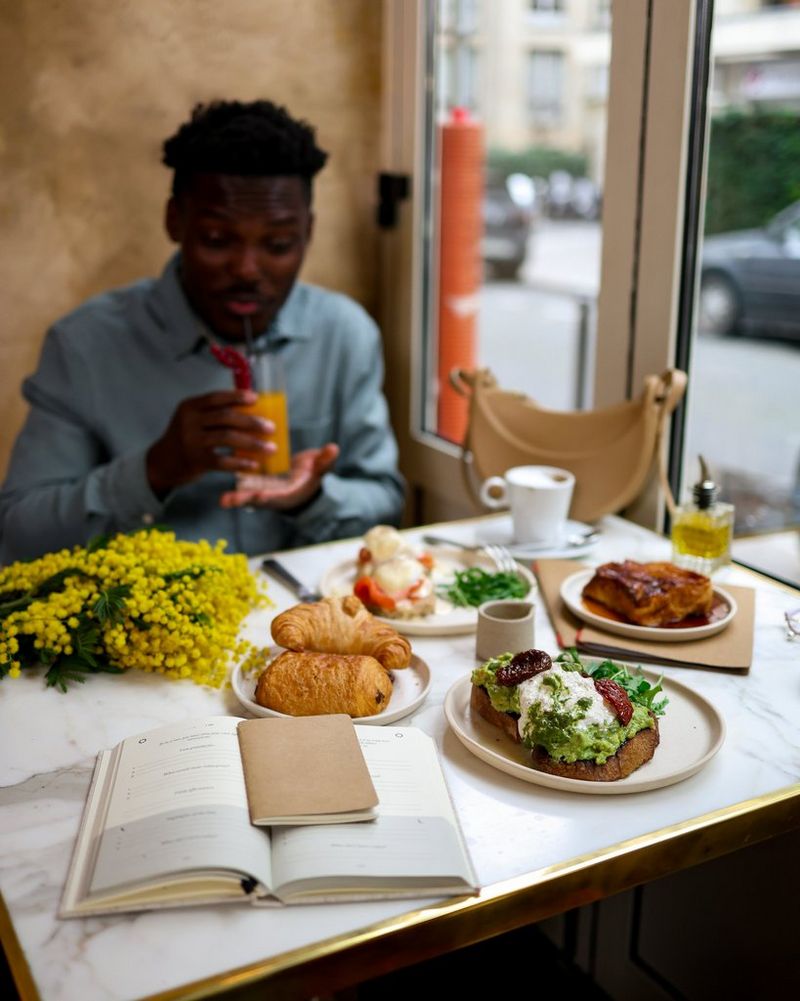 Tomi Adebayo sits at a restaurant table covered in plates of food, drinking from a glass with a straw, in a photograph taken by Laura Hannoun.
