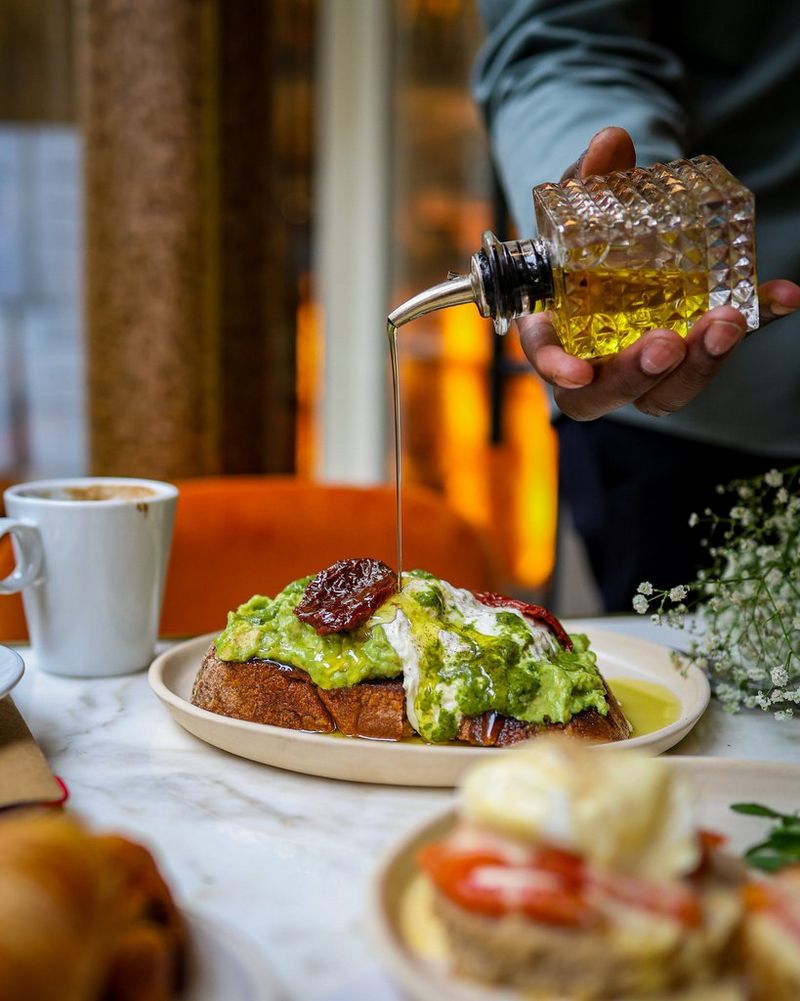 A hand pours oil over a plate of avocado on toast garnished with sundried tomatoes, in a photograph taken by Laura Hannou