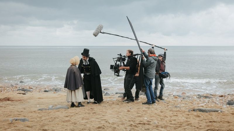 A camera crew filming two actors beside the sea: a Victorian gentleman in a top hat and cape leaning over to talk to a young girl.
