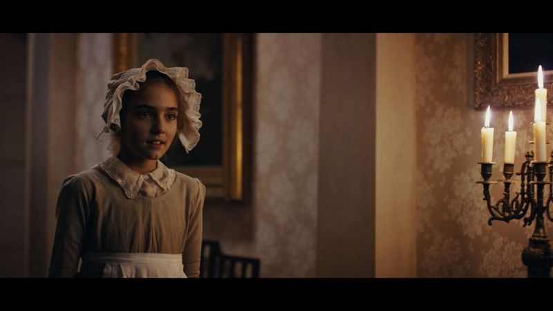 Kiara Holley-Paliano as Mary Anning in Regency dress in an interior manor set with candles to one side.