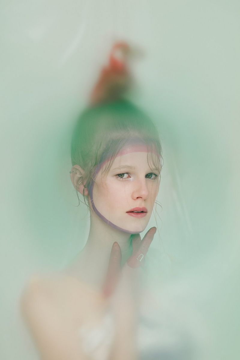 Le visage d'une personne apparaît au premier plan, tandis que le reste de sa tête (ainsi que l'oiseau rouge perché à son sommet) et ses épaules sont floues, comme dans un smog aux couleurs pastel. Photographie de Laura Knipsael.