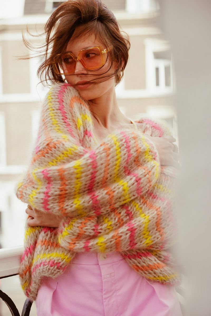 Une personne portant des lunettes de soleil pose sur un balcon, serrant autour d'elle un tricot couleur pastel. Photographie prise avec un Canon EOS R5 par Laura Knipsael.