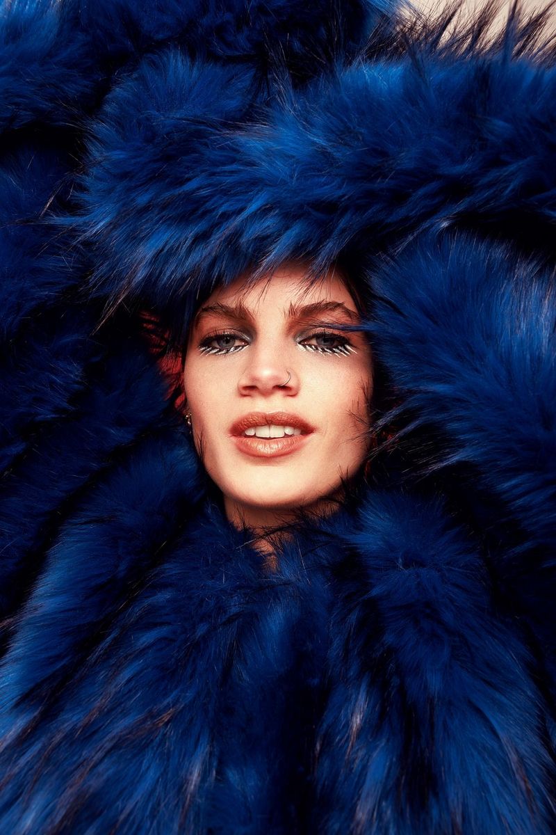 Une personne tient ses bras au-dessus de sa tête, vêtue d'un manteau bleu ressemblant à de la fourrure, son visage entouré par le tissu au centre. Photographie prise avec un Canon EOS R5 par Laura Knipsael.