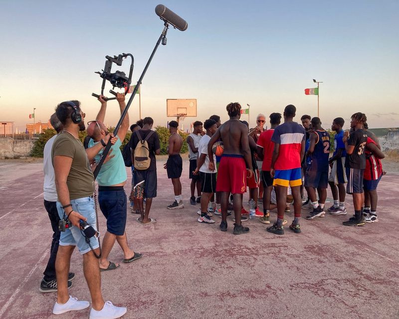 Un gruppo di giocatori di basket si riunisce sul campo intorno al loro allenatore. Vengono ripresi da tre filmmaker con videocamera e microfono che si trovano lì vicino.