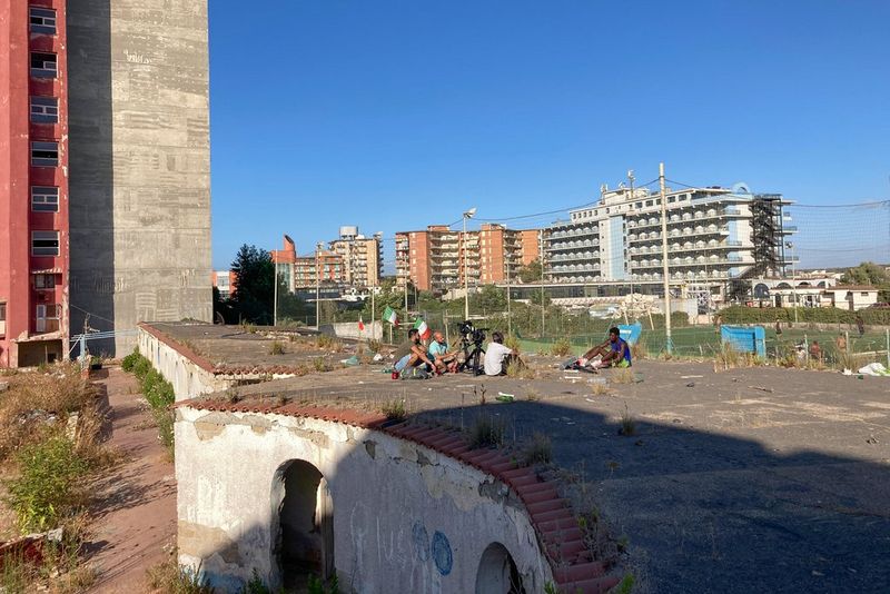 Un gruppo di persone è seduto sul tetto di un edificio abbandonato. Tre di loro puntano due videocamere su treppiedi al quarto, che viene intervistato.