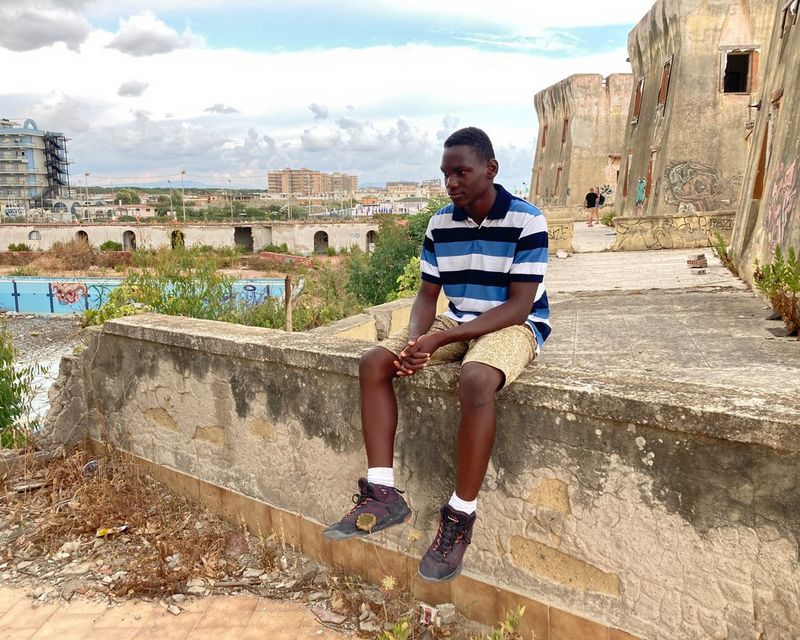 Un adolescente con una polo a righe e dei pantaloncini corti è seduto su un muretto scrostato con le mani giunte, sullo sfondo sono visibili degli edifici abbandonati.