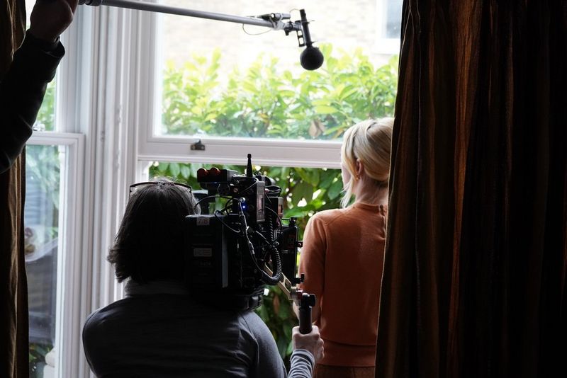Cinematographer Tania Freimuth is pictured from behind filming actress Eloise Smyth looking out of a window. 