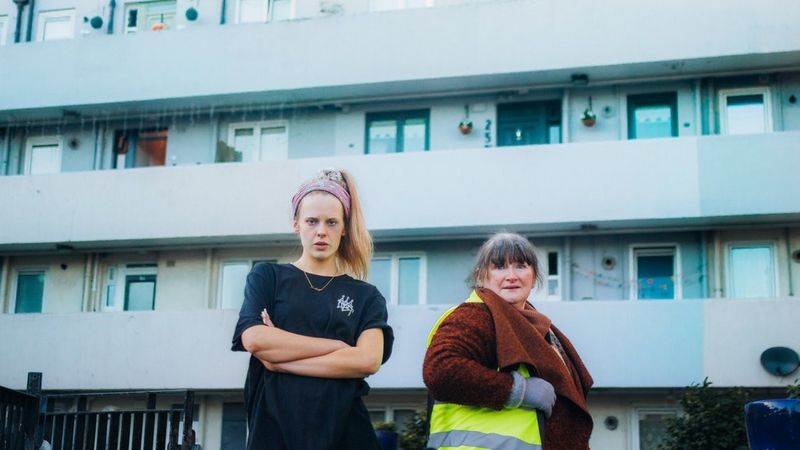 A young woman and a middle-aged woman stand in front of a block of flats, facing towards the camera, in a publicity photo from the film Verdigris. Photo credit: Eleanor Rogers