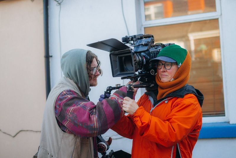 Cinematographer Tania Freimuth, wearing a bright orange raincoat, stands outside holding a camera rig with a Canon EOS C500 Mark II camera on it. An assistant leans towards her to adjust the rigging.