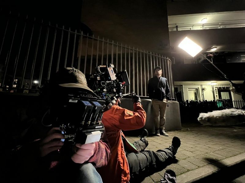 Cinematographer Tania Freimuth sits on the pavement, pointing her camera up at a man standing in front of some railings. The scene is at night with a light being held at the side.