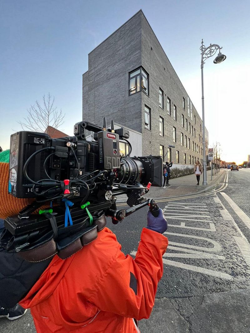 Cinematographer Tania Freimuth, wearing a bright orange raincoat and carrying a camera rig holding a Canon EOS C500 Mark II, faces across a road and towards a pavement by the side of a large grey building.