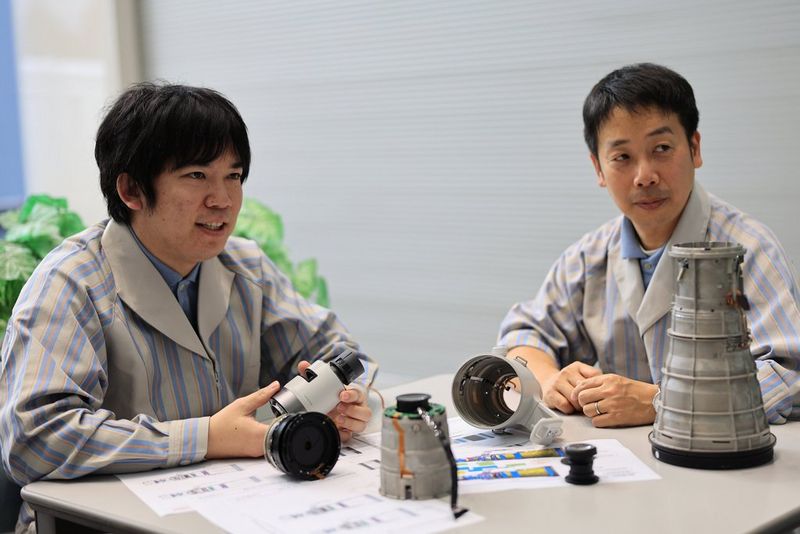 Deux hommes vêtus de blouses de laboratoire rayées sont assis à un bureau. Des éléments d'un super téléobjectif Canon sont posés sur la table devant eux.