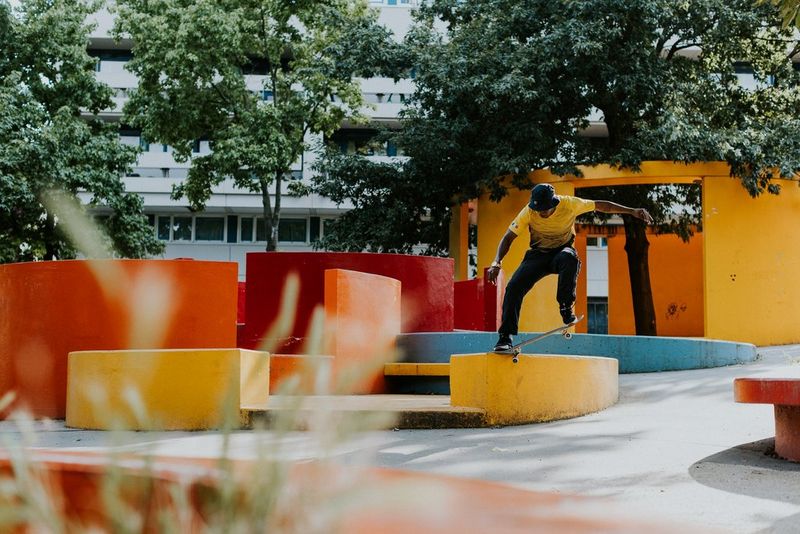 Un uomo esegue un trick su uno skateboard con uno skatepark pieno di colori sullo sfondo. Scatto realizzato con Canon EOS R5 da Teddy Morellec.
