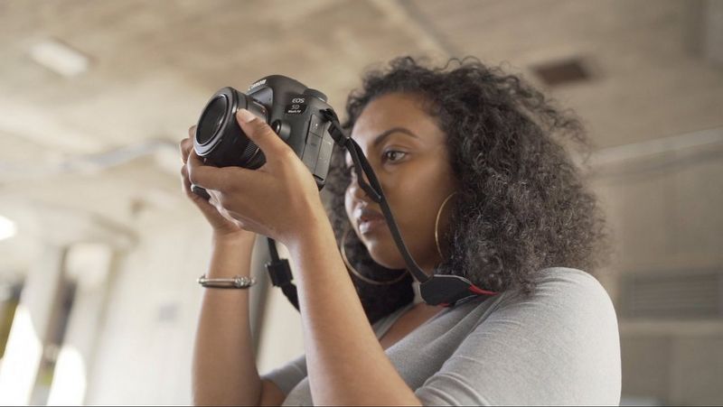 Видеограф Лорен Макколин в сером топе и с большими серьгами-кольцами снимает на камеру Canon с высоты плеч.