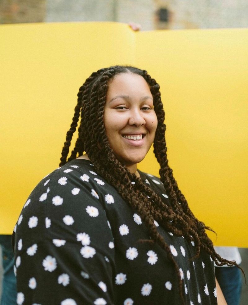 Видеограф Эмбер Акауну улыбается на камеру на ярко-желтом фоне.