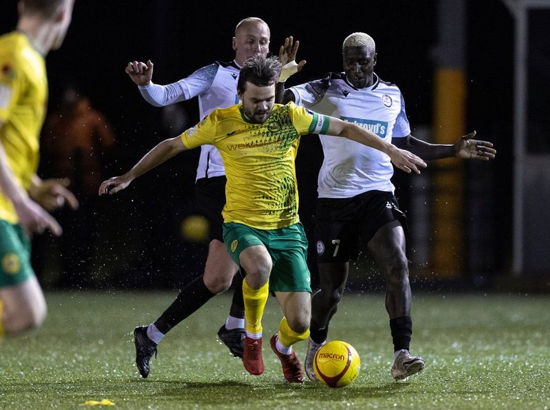 In a photo taken on a Canon EOS R3 by Eddie Keogh under weak floodlights, a footballer in a yellow jersey dribbles the ball at his feet while two footballers in white jerseys run behind him.