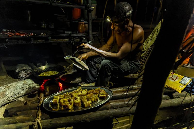 In a photo taken on a Canon EOS R3 by Christian Ziegler in low light conditions, a shirtless man wearing a head torch holds a ladle while food cooks in a wok on a fire in front of him.