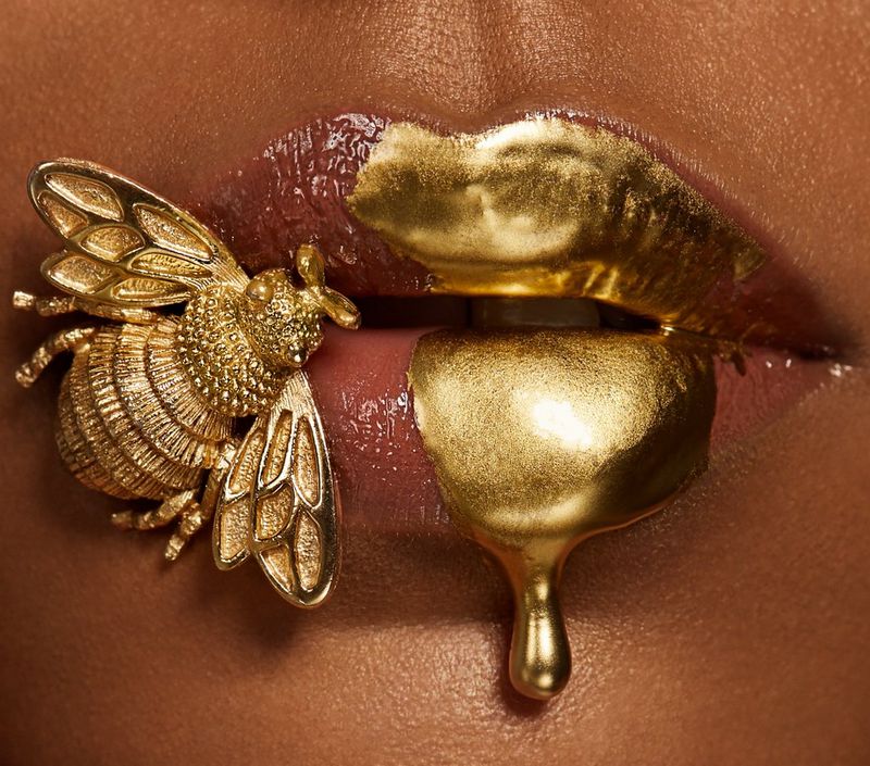 A gold metal bee sits on a female model's made-up lips that are dripping with gold paint.