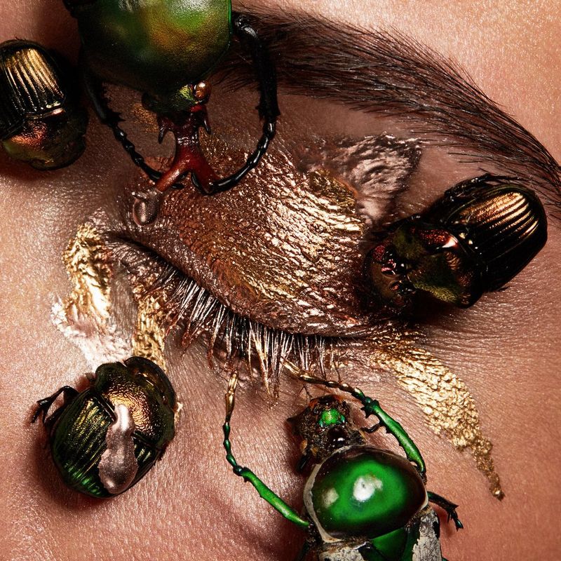 Iridescent beetles sit on the skin around a closed eyelid, which is smeared in gold eyeshadow.