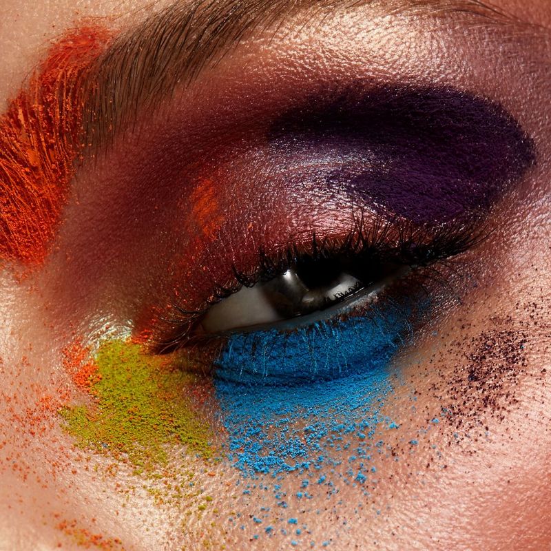 An open eye stares at the camera, the eyelid and surrounding skin covered in different coloured powders including blue, red, yellow and purple.