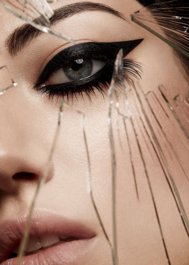 A model's eye, heavily made up with sculpted black eyeliner, looks through a gap in shards of broken glass.