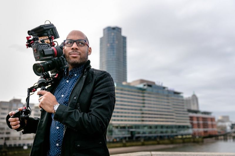 A man with a Canon Cinema EOS camera on his shoulder by a riverfront.