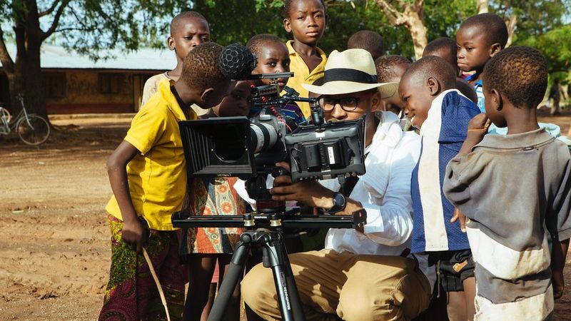 A man filming with a Canon EOS Cinema camera, surrounded by children looking at the equipment.