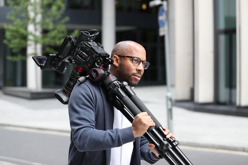 A man holding a Cinema EOS camera on a tripod over his shoulder on a quiet street.
