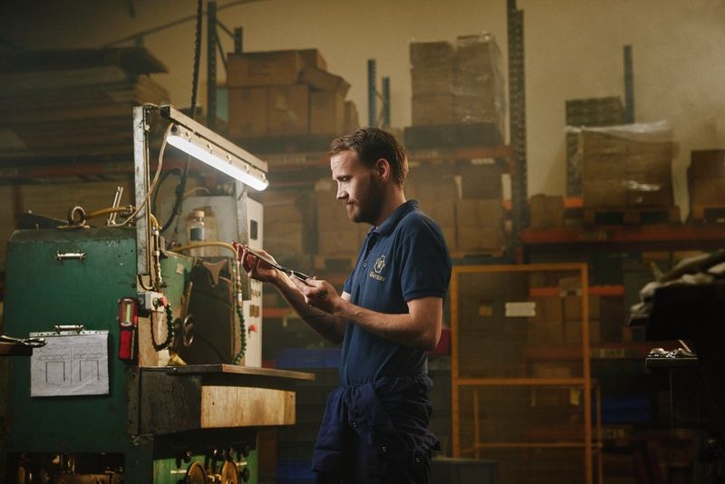 A man stands in a workshop against a bench, holding and looking at one half of a pair of scissors, taken on a Canon EOS R5 C by Tom Barnes.