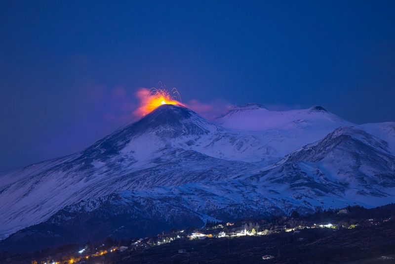 A night shot taken on a Canon EOS R5 by travel photographer Ulla Lohmann of a volcano spouting fiery red, looming over the lights of a village stretching across its foot.