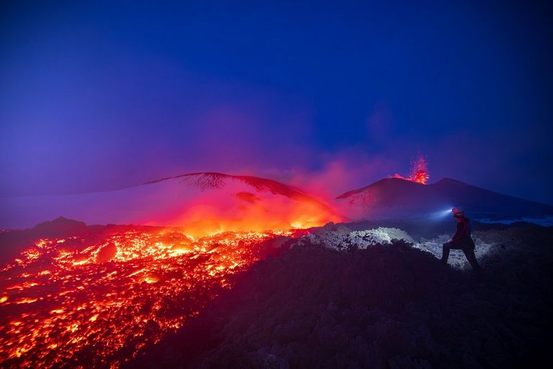 A small figure with a head torch stands just next to a glowing red lava flow, with an erupting volcano in the background. Adventure travel photo by Ulla Lohmann taken on a Canon EOS R5.