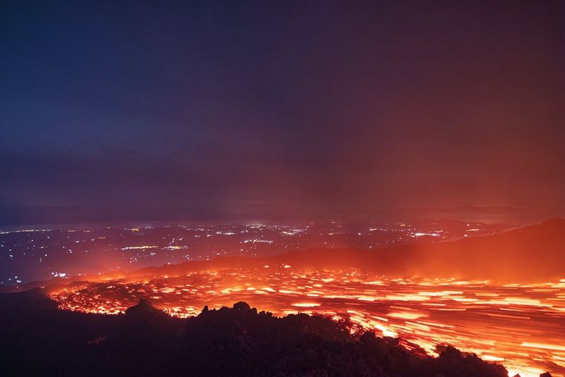 A field of molten lava glows in the darkness, with the lights of human settlements in the distant background. Photo taken by Ulla Lohmann on a Canon EOS R5.