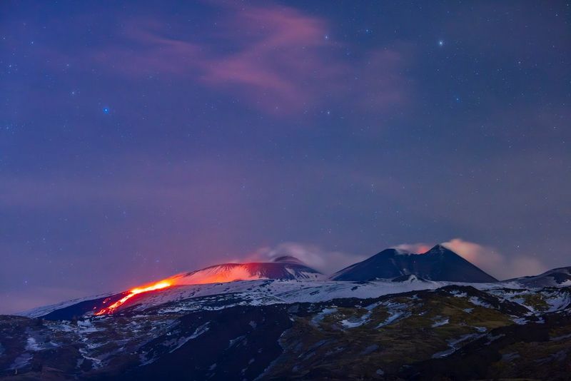 A lava flow glows red in an evening mountain landscape, with a smoking volcano in the background and stars in the sky, photographed by Ulla Lohmann on a Canon EOS R5.