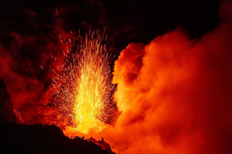 An adventure travel photo by Ulla Lohmann taken on a Canon EOS R5 captures a fiery red and orange volcanic eruption, silhouetting tiny people observing it in the foreground.