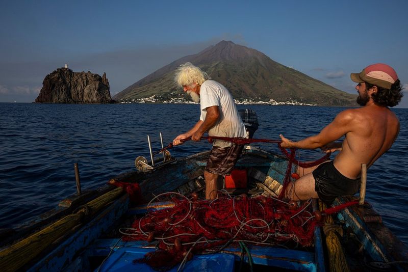A young fisherman and an older one haul in their net with a smoking volcano rising out of the sea in the background. Travel photo by Ulla Lohman taken on a Canon EOS R5 Mark II.