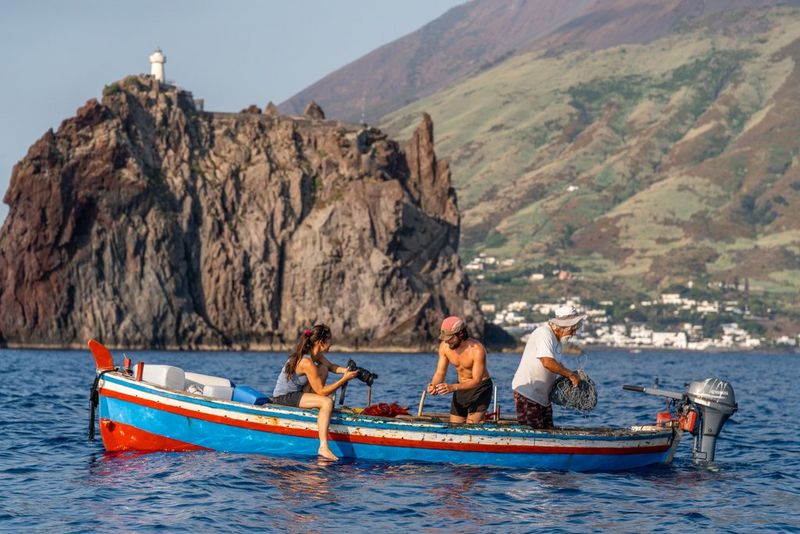 Adventure photographer and filmmaker Ulla Lohmann sits with her camera in s small fishing boat with two fishermen at work, with a craggy volcanic coastline in the background.