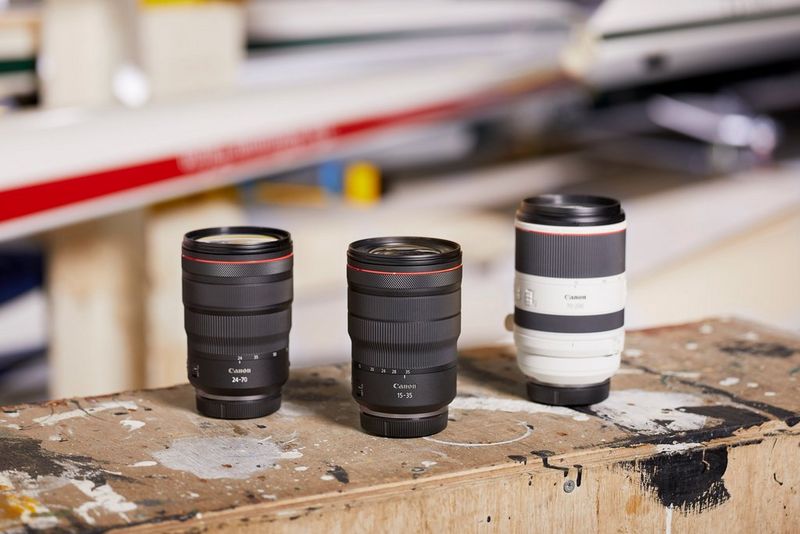 Three Canon RF zoom lenses, sitting on a paint-spattered wooden surface. A rowing scull with a white-and-red hull is out of focus in the background.