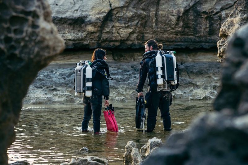 Due figure in muta da sub sono in piedi nell'acqua bassa, circondati da formazioni rocciose.
