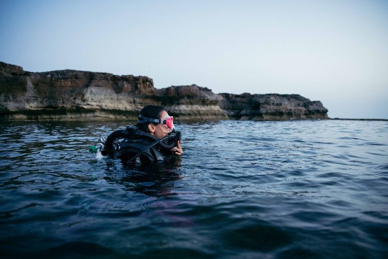 La testa e le spalle di un sub emergono dall'acqua sullo sfondo di una costa rocciosa.